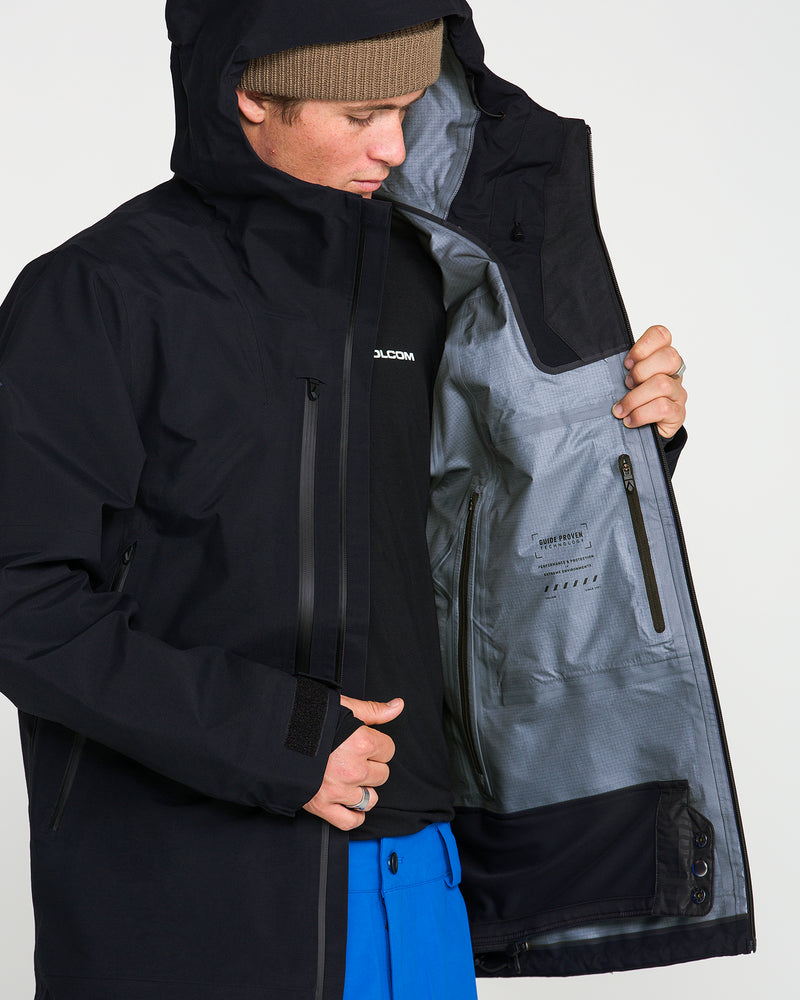 A person wearing the Volcom Men’s Guch 3L Gore-Tex Pro Jacket in black recycled-nylon shell, with the hood up and paired with blue pants, opens one side to reveal a gray inner lining featuring a zippered pocket—perfect for stashing essentials on backcountry adventures.
