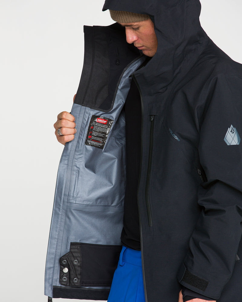 A person wears the Volcom Men’s Guch 3L GORE-TEX Pro Jacket, paired with blue pants and a brown beanie, looking down while holding open the jacket to reveal its gray inner lining and mesh pocket featuring a red-tagged label—showcasing the premium recycled-nylon shell engineered for backcountry adventures.