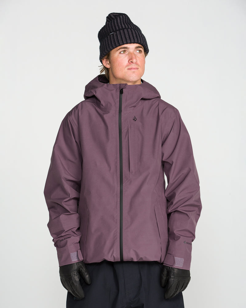 A person wearing the Volcom Unisex Tester 3L Gore-Tex Jacket—a dark purple hooded jacket made from PFAS-free, recycled 3-layer fabric—along with a black beanie and black gloves, stands against a plain light background, looking up and to the side.