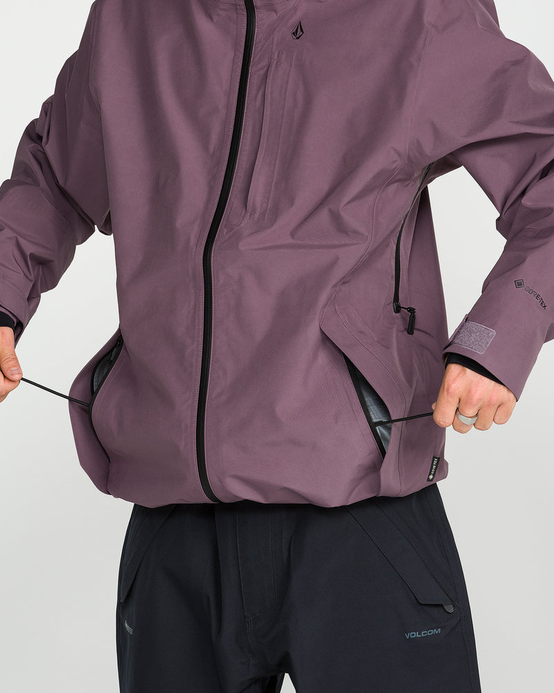 A person adjusts the drawstrings on a Unisex Tester 3L Gore-Tex Jacket by Volcom, featuring a purple hue with black zippers and paired with black pants. Made from recycled 3-layer fabric, this jacket combines durable construction with eco-conscious design. Only the torso and hands are visible.