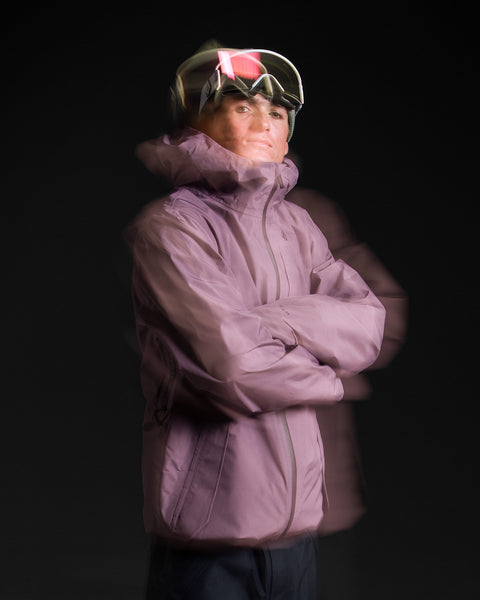 A person wearing the Volcom Unisex Tester 3L Gore-Tex Jacket in purple and ski goggles stands with arms crossed against a dark background. A motion blur effect adds a sense of movement to the image.