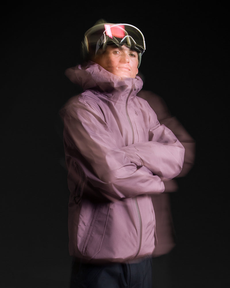 A person wearing the Volcom Unisex Tester 3L Gore-Tex Jacket in purple and ski goggles stands with arms crossed against a dark background. A motion blur effect adds a sense of movement to the image.