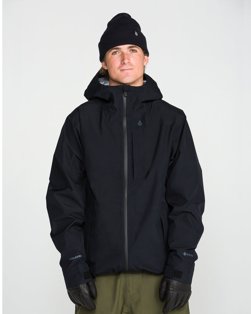 A person wearing the Volcom Unisex Tester 3L Gore-Tex Jacket in black, paired with a black beanie and black gloves, stands against a plain white background, looking slightly upward.