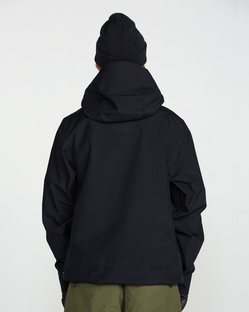A person is standing with their back to the camera against a plain white background, wearing a black Volcom Unisex Tester 3L Gore-Tex Jacket with a hood, a black knit beanie, and green pants.