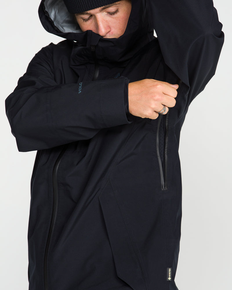 A person wearing a black Volcom Unisex Tester 3L Gore-Tex Jacket, which is waterproof and PFAS-free, zips a pocket on their upper arm while facing slightly downward. They also have on a black beanie.