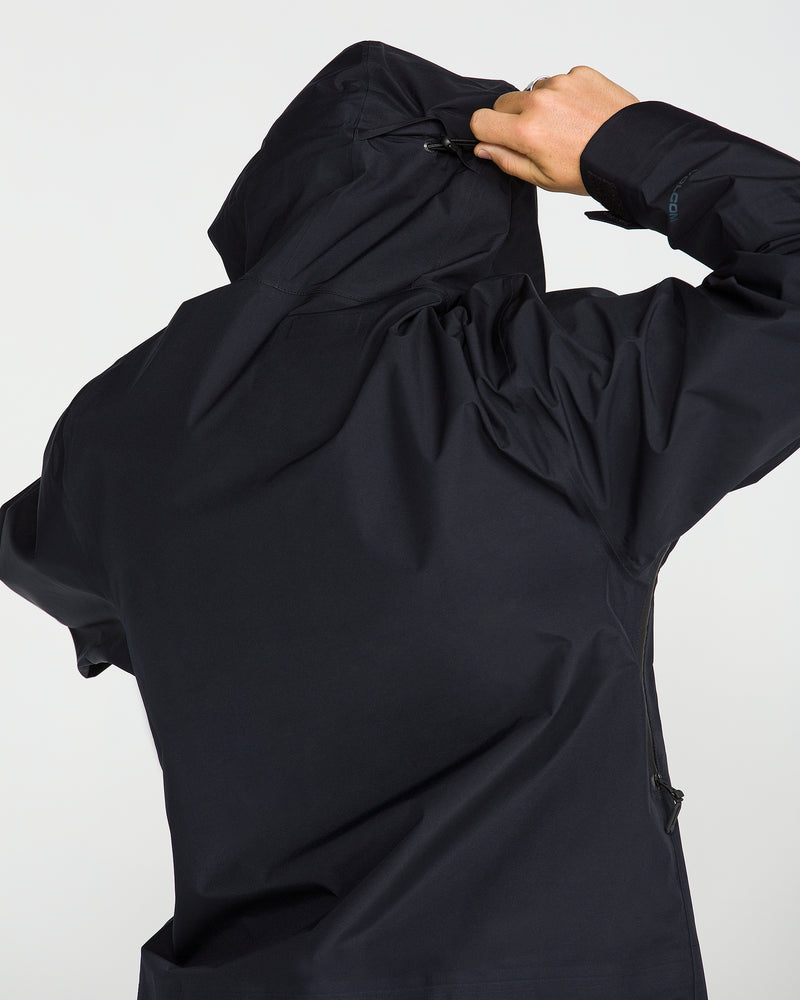 A person wearing the Volcom Unisex Tester 3L Gore-Tex Jacket in black is seen from behind, adjusting the hood with one hand against a plain light background.