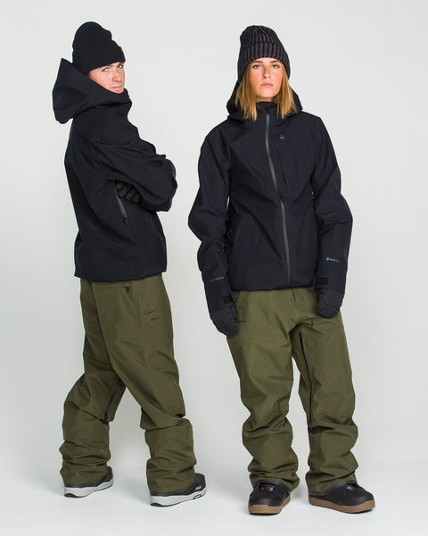 Two people stand against a plain white background, each wearing the Volcom Unisex Tester 3L GORE-TEX Jacket in black, paired with olive green snow pants, black gloves, and black beanies. Both are wearing snow boots and have a relaxed posture. The jackets feature a PFAS-free design for environmentally conscious protection from the elements.