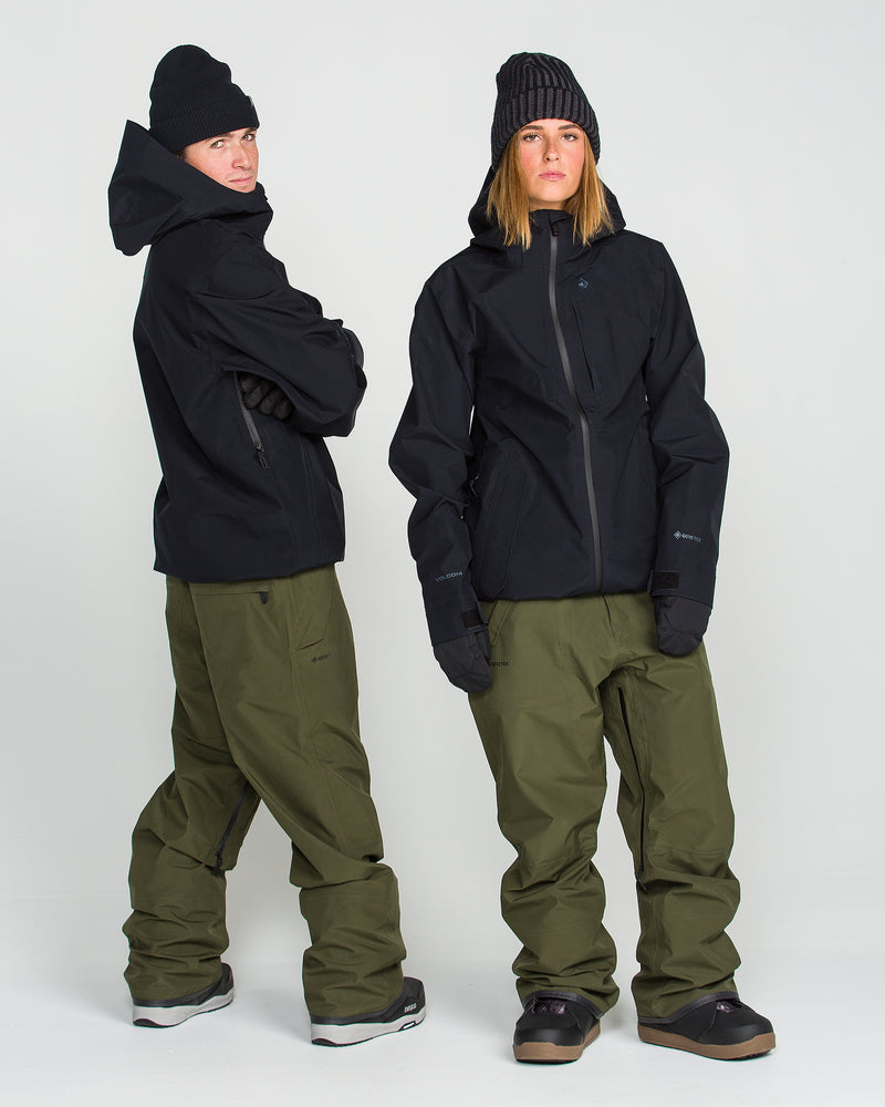 Two people stand against a plain white background, each wearing the Volcom Unisex Tester 3L GORE-TEX Jacket in black, paired with olive green snow pants, black gloves, and black beanies. Both are wearing snow boots and have a relaxed posture. The jackets feature a PFAS-free design for environmentally conscious protection from the elements.