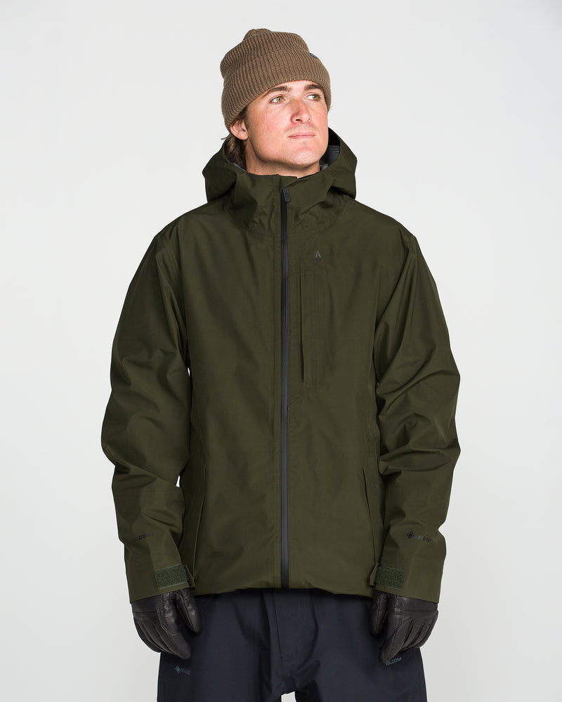 A person wearing a brown beanie, black gloves, dark pants, and the Volcom Men’s Tester 3L Gore-Tex Jacket in dark green—crafted from recycled fabric—stands against a plain white background, looking slightly up and to the side.