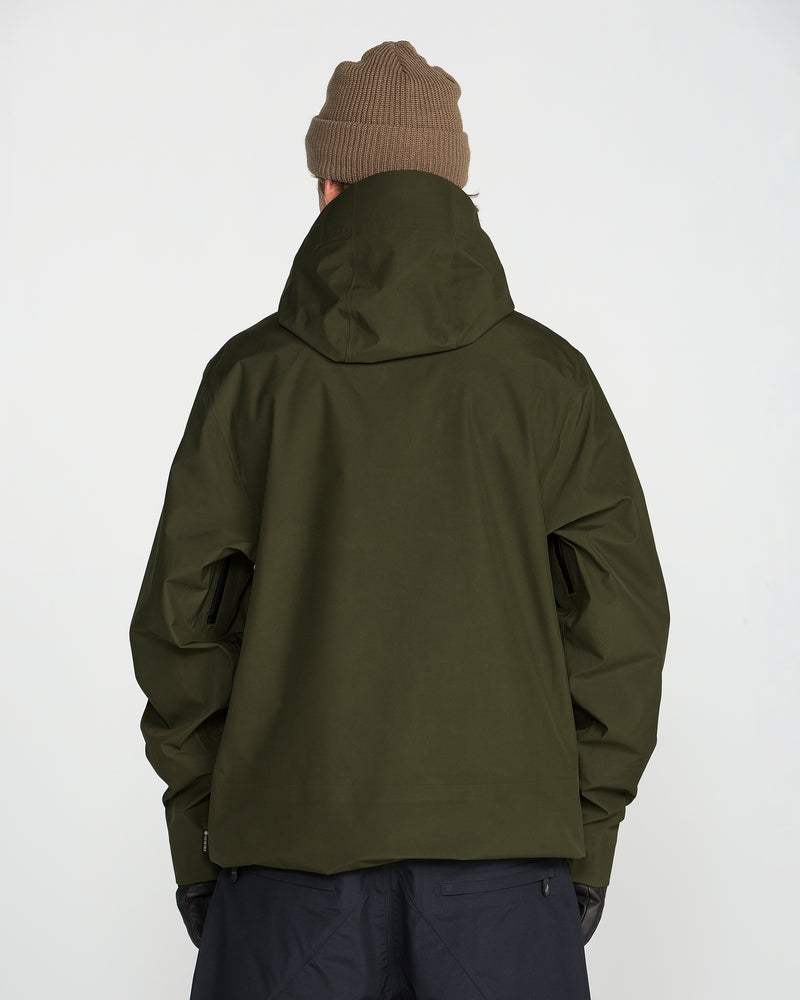 A person wearing a brown beanie, black pants, and the Volcom Men’s Tester 3L Gore-Tex Jacket in olive green stands facing away against a plain white background.