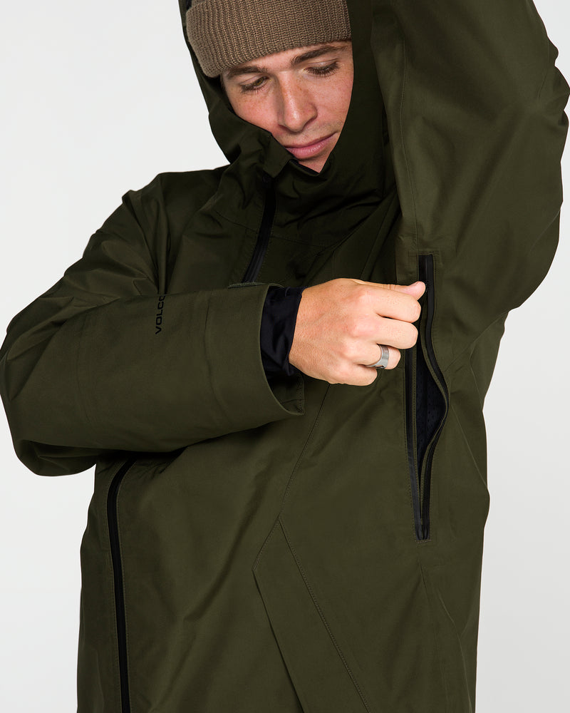 A man wearing the Volcom Men’s Tester 3L Gore-Tex Jacket in olive green—crafted from recycled fabric—opens a zippered underarm vent while looking down to adjust the garment, with a brown beanie completing his look.