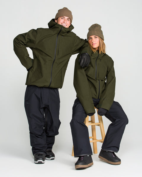 Two people in Volcom Men’s Tester 3L Gore-Tex Jackets pose in a studio, each styled with black pants, boots, and brown beanies. One stands confidently with a hand on their hip, while the other sits on a wooden stool with one foot resting on the ground.