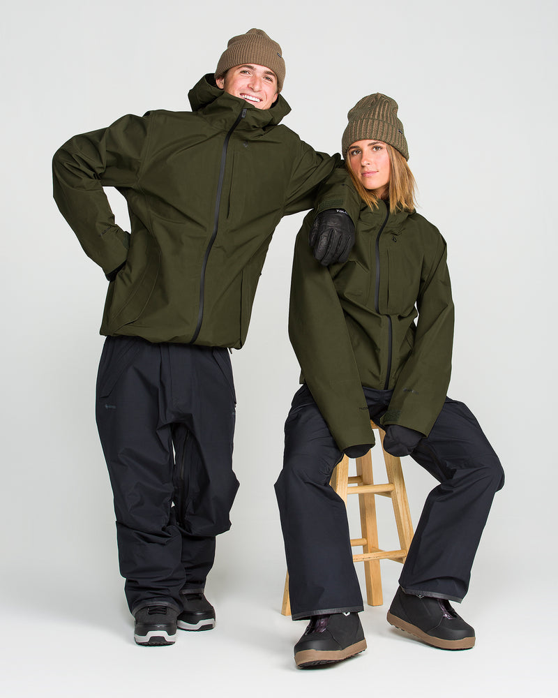 Two people in Volcom Men’s Tester 3L Gore-Tex Jackets pose in a studio, each styled with black pants, boots, and brown beanies. One stands confidently with a hand on their hip, while the other sits on a wooden stool with one foot resting on the ground.