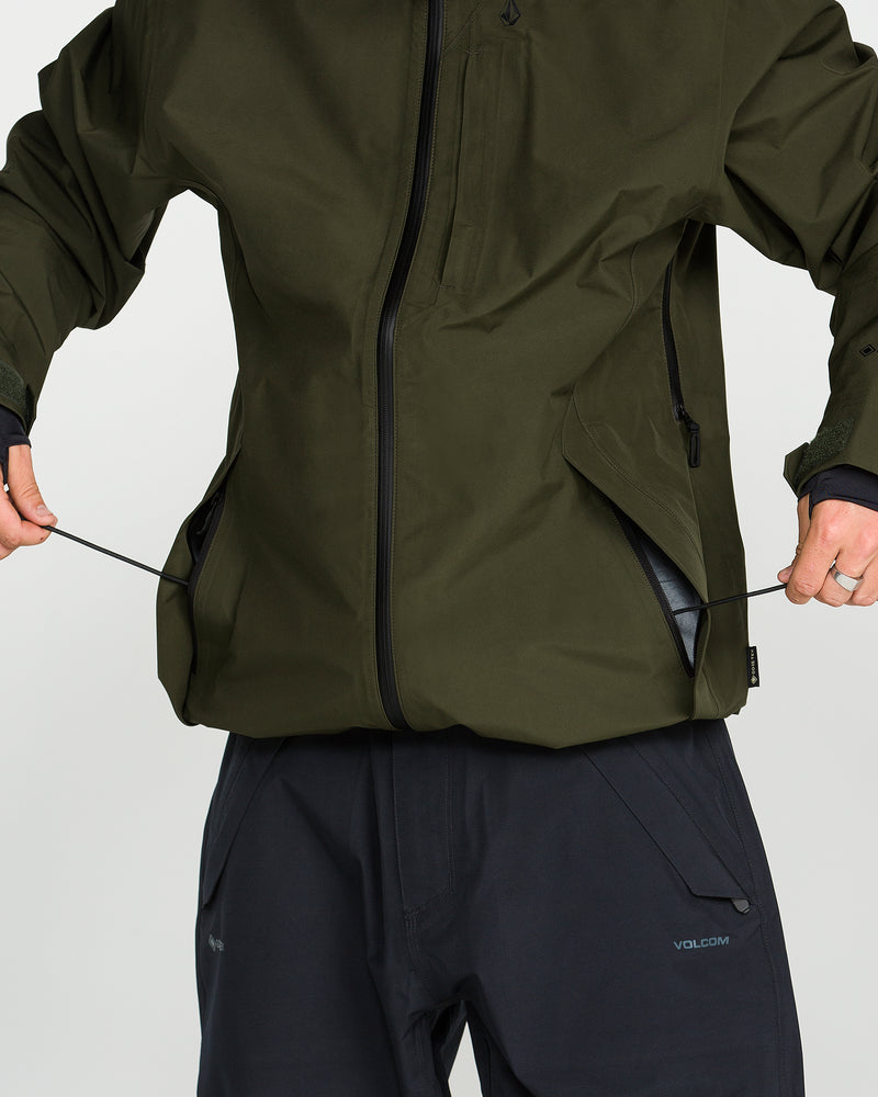 A person wearing the Volcom Men’s Tester 3L Gore-Tex Jacket in olive green, made from recycled fabric, pairs it with black pants and adjusts the jacket’s hem by pulling its drawstrings. Only their torso and hands are visible.