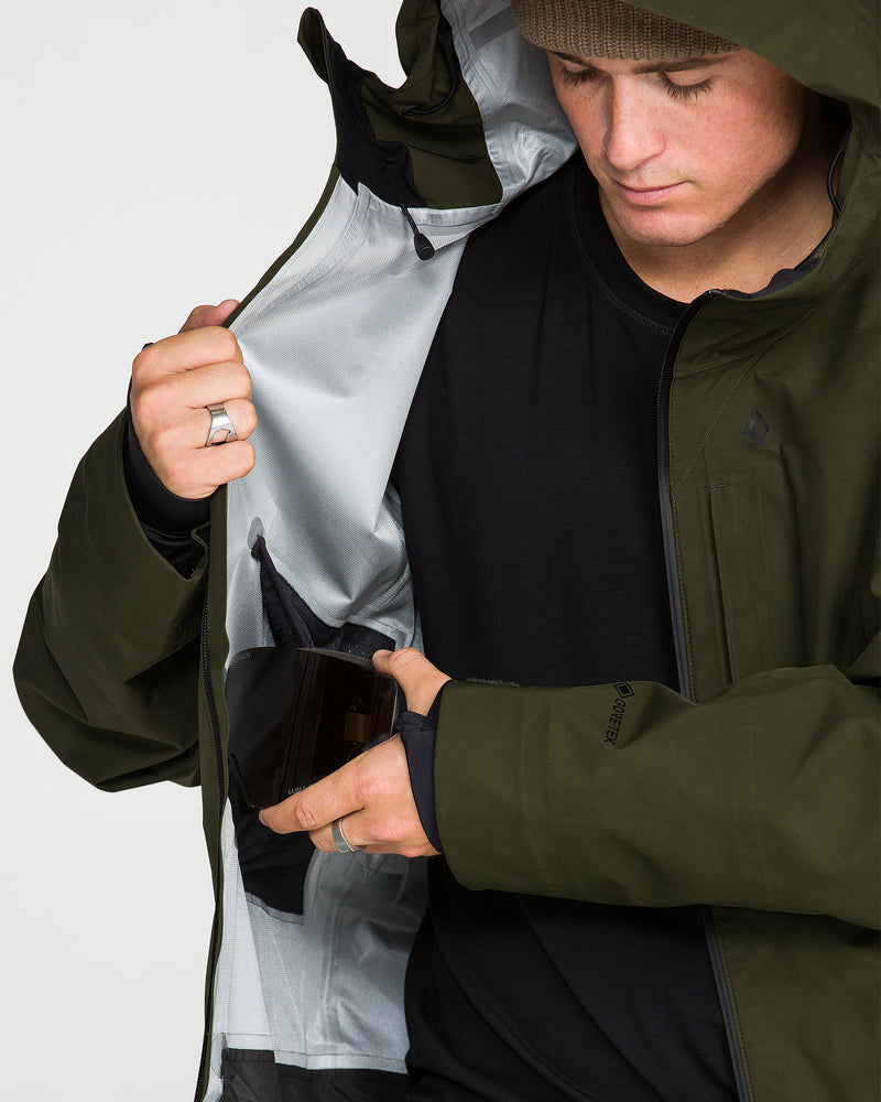 Wearing a green Volcom Men’s Tester 3L Gore-Tex Jacket and a brown beanie, a person places a wallet into an inner jacket pocket while looking down.
