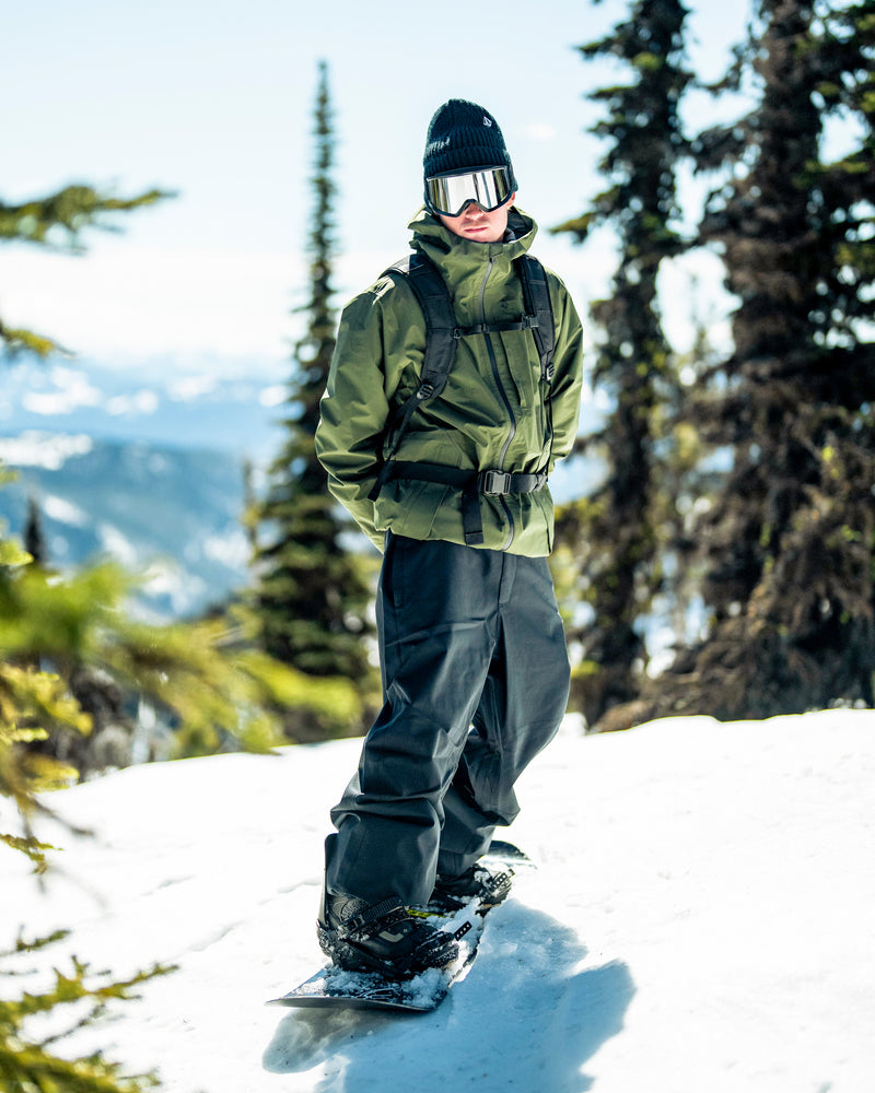 A person wearing the Volcom Unisex Tester 3L Gore-Tex Jacket, along with black pants, gloves, and goggles, stands on a snowboard in a snowy mountain landscape with trees and distant mountains in the background.