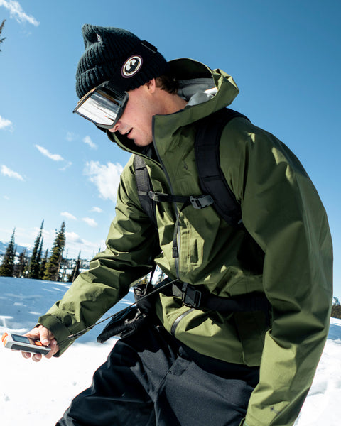 A person wearing the Volcom Unisex Tester 3L Gore-Tex Jacket, along with a black backpack, black beanie, and ski goggles, uses a handheld device in the snow on a sunny day, with trees and a blue sky in the background.