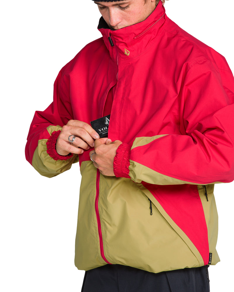 A person wearing a red and tan Men’s Longo Gore-Tex Jacket, pulling a black tag from the chest pocket.