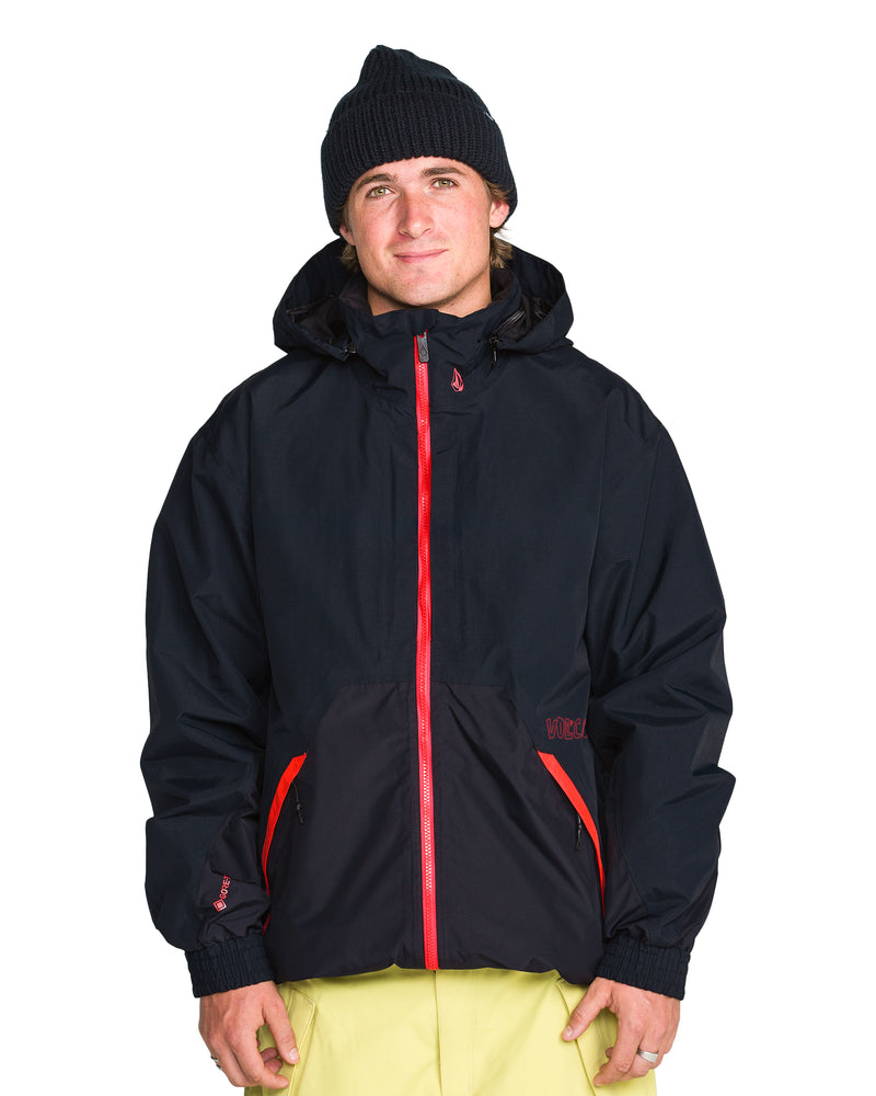 Man wearing a dark hooded Volcom Men’s Longo Gore-Tex Jacket with a bright red zipper and yellow pants.