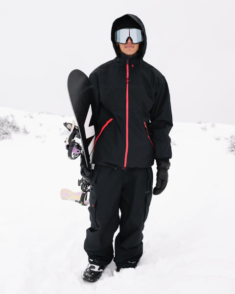 A person wearing the Volcom Men’s Longo GORE-TEX Jacket along with snow goggles and gloves stands in the snow, holding a snowboard upright on their left side against an overcast, snowy background.