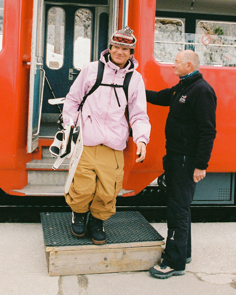 A person in a pink snowboard jacket and tan pants steps off a red train carrying a snowboard, while another person stands nearby, smiling and wearing the Volcom Men’s Longo Gore-Tex Jacket.