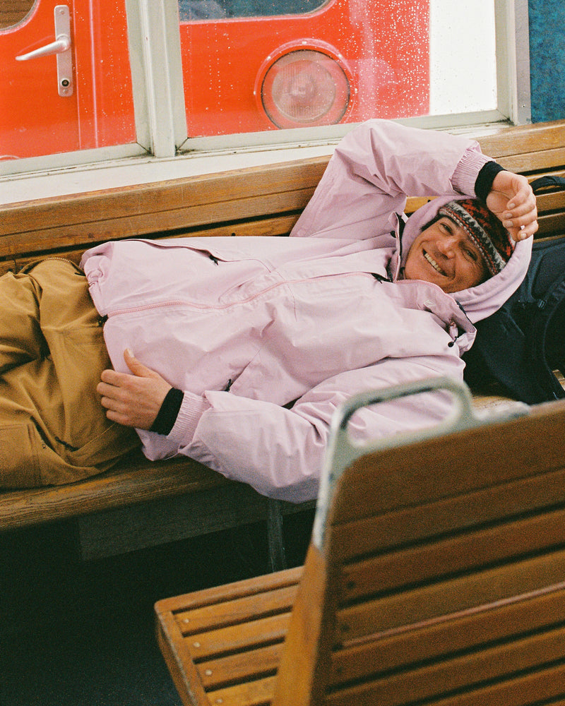 Wearing the Volcom Men’s Longo Gore-Tex Jacket in pink and tan pants, a person lies on a wooden bench, smiling as they shield their eyes from the light. A red vehicle or structure can be seen through the window behind them.