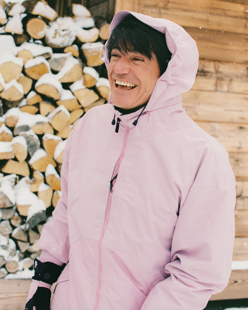 Wearing the Volcom Men’s Longo Gore-Tex Jacket in light pink, a person smiles outdoors near a stacked woodpile, with a wooden building and snow in the background.