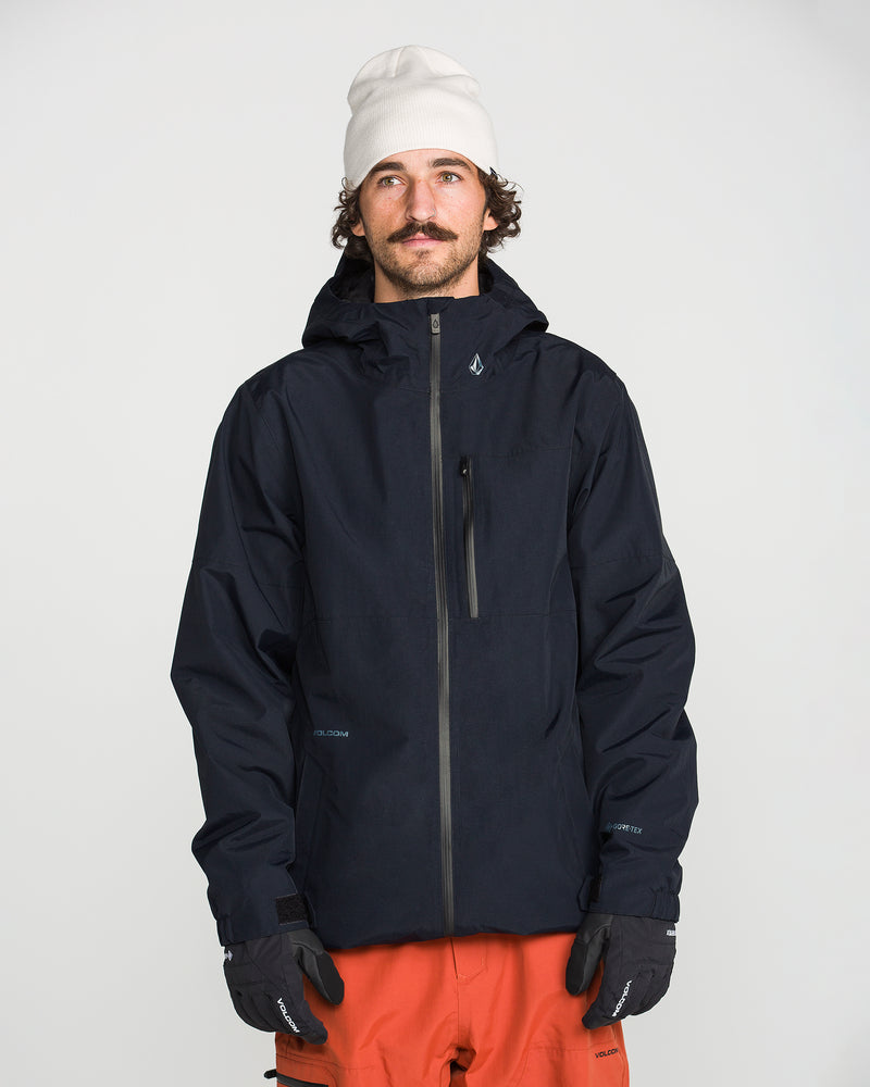 A man with a mustache wears a white beanie, black Volcom Men’s L Gore-Tex Jacket with a recycled shell, black gloves, and orange pants, standing against a plain white background and looking slightly upward.