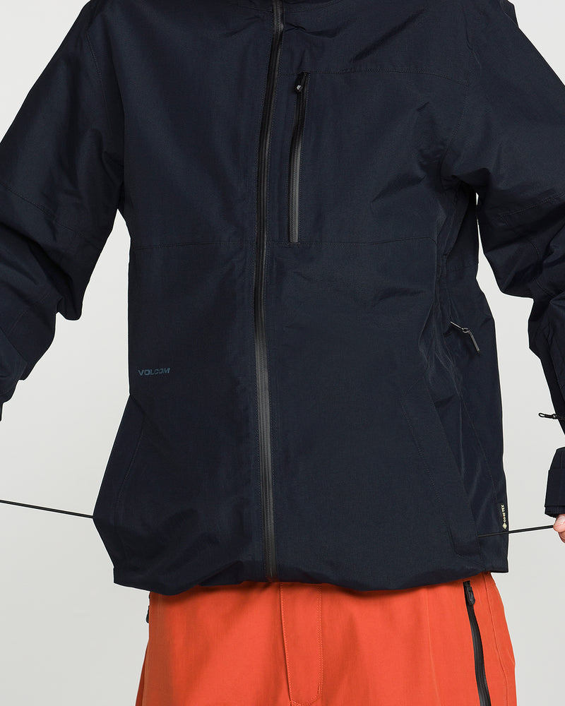 A person is shown from the shoulders to the thighs against a plain background, wearing a Volcom Men’s L Gore-Tex Jacket with an unzipped front and recycled shell in black, paired with bright orange pants.