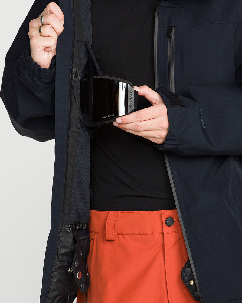 Wearing the Volcom Men’s L Gore-Tex Jacket and orange pants, a person places ski goggles into an inner pocket, highlighting the jacket’s waterproof performance and well-considered design.