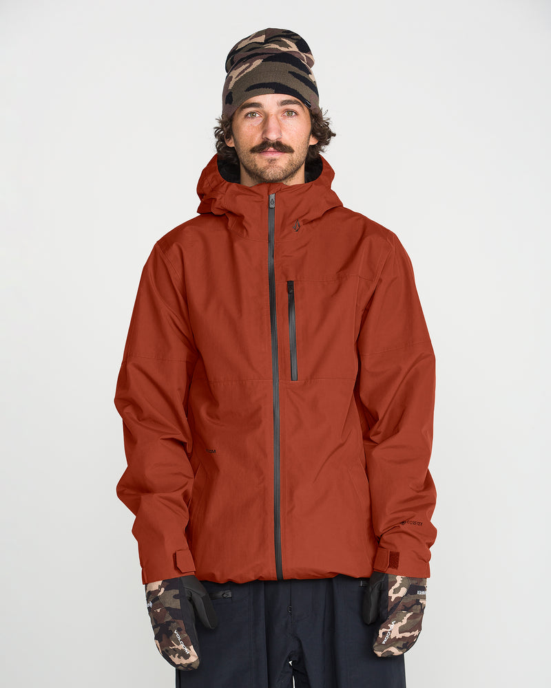 A person wearing a rust-colored Volcom Men’s L Gore-Tex Jacket, paired with a patterned beanie and camouflage gloves, stands facing forward against a plain white background.