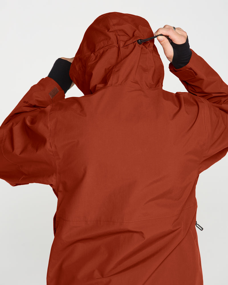 A person wearing the Volcom Men’s L Gore-Tex Jacket in rust is seen from behind, adjusting the drawstring on the hood. Made from recycled nylon, this waterproof jacket stands out against a plain, light-colored background.