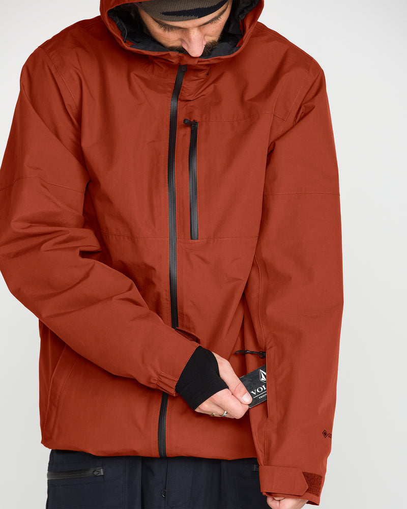 A person wearing a rust-colored Volcom Men’s L Gore-Tex Jacket made from recycled nylon unzips a small pocket on the left sleeve, revealing a card or pass.