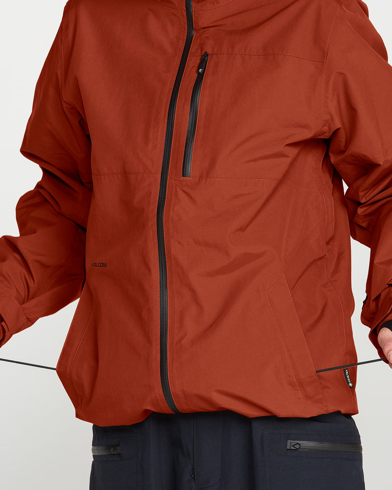 A person is shown from the shoulders to the upper legs against a plain background, wearing Volcom’s Men’s L Gore-Tex Jacket in rust with black zippers and dark pants.