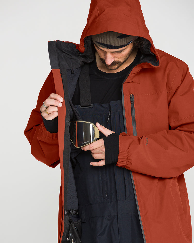 Wearing the rust-colored Volcom Men’s L Gore-Tex Jacket, a person opens it to reveal black overalls and stores ski goggles inside.