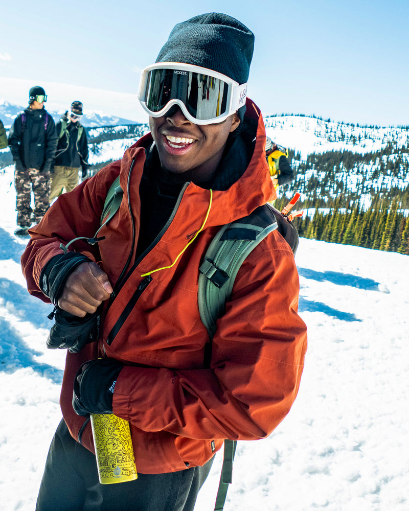 A person in a vibrant orange Volcom Men’s L Gore-Tex Jacket, black gloves, and white ski goggles smiles while holding a yellow can on a snowy mountain with others standing in the background.