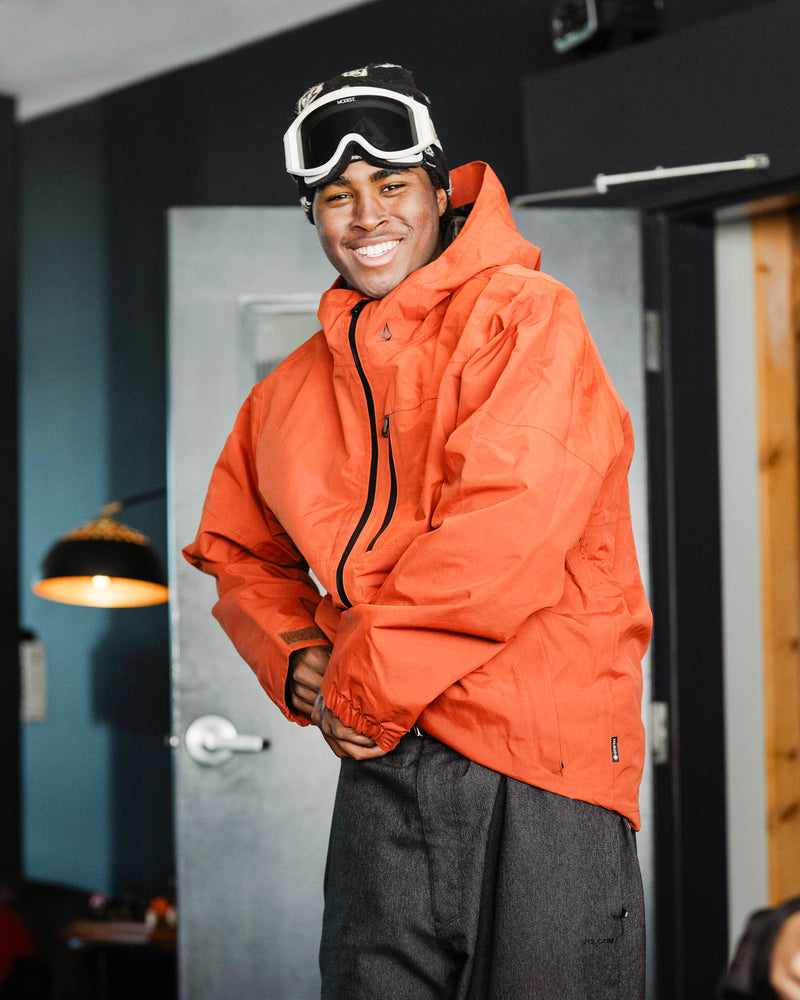A person wearing a Volcom Men’s L Gore-Tex Jacket in orange and ski goggles on their head stands indoors, smiling and posing with hands clasped in front.