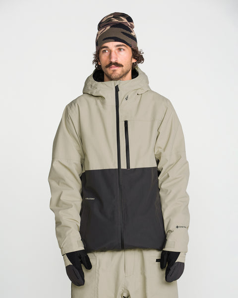 A person wearing a tan and black Volcom Men’s L Gore-Tex Jacket, tan pants, black gloves, and a patterned beanie stands against a plain white background.