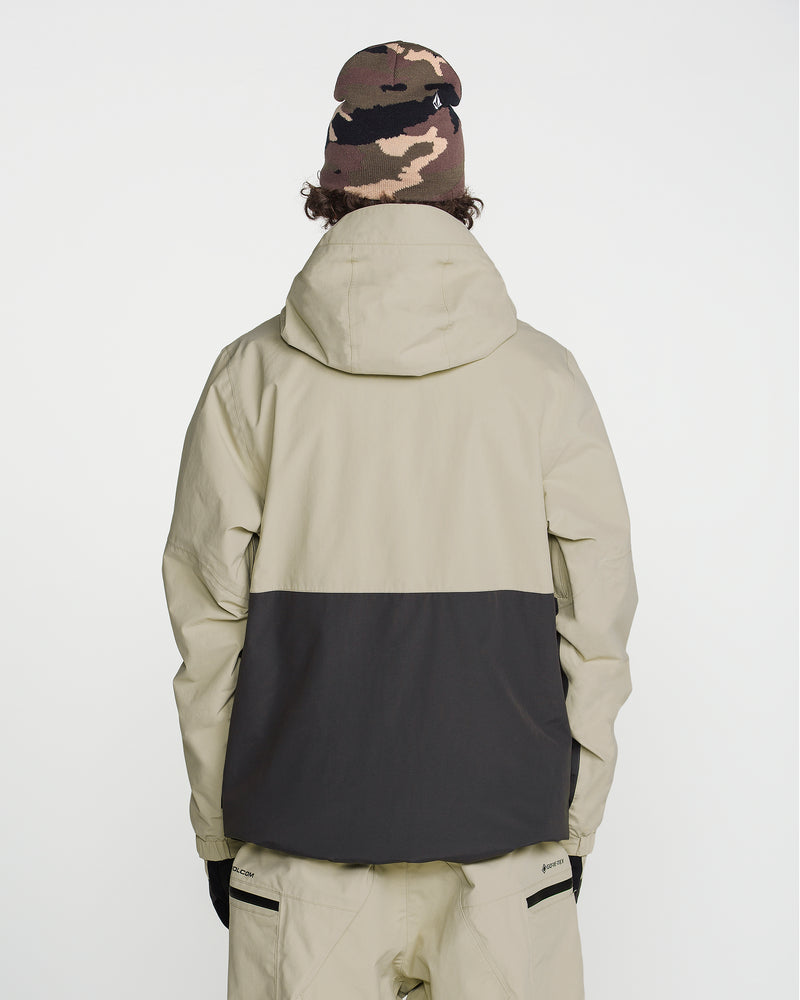 A person facing away from the camera wears a Volcom Men’s L Gore-Tex Jacket in beige and black, along with beige pants, black gloves, and a brown camouflage beanie. The background is plain white.