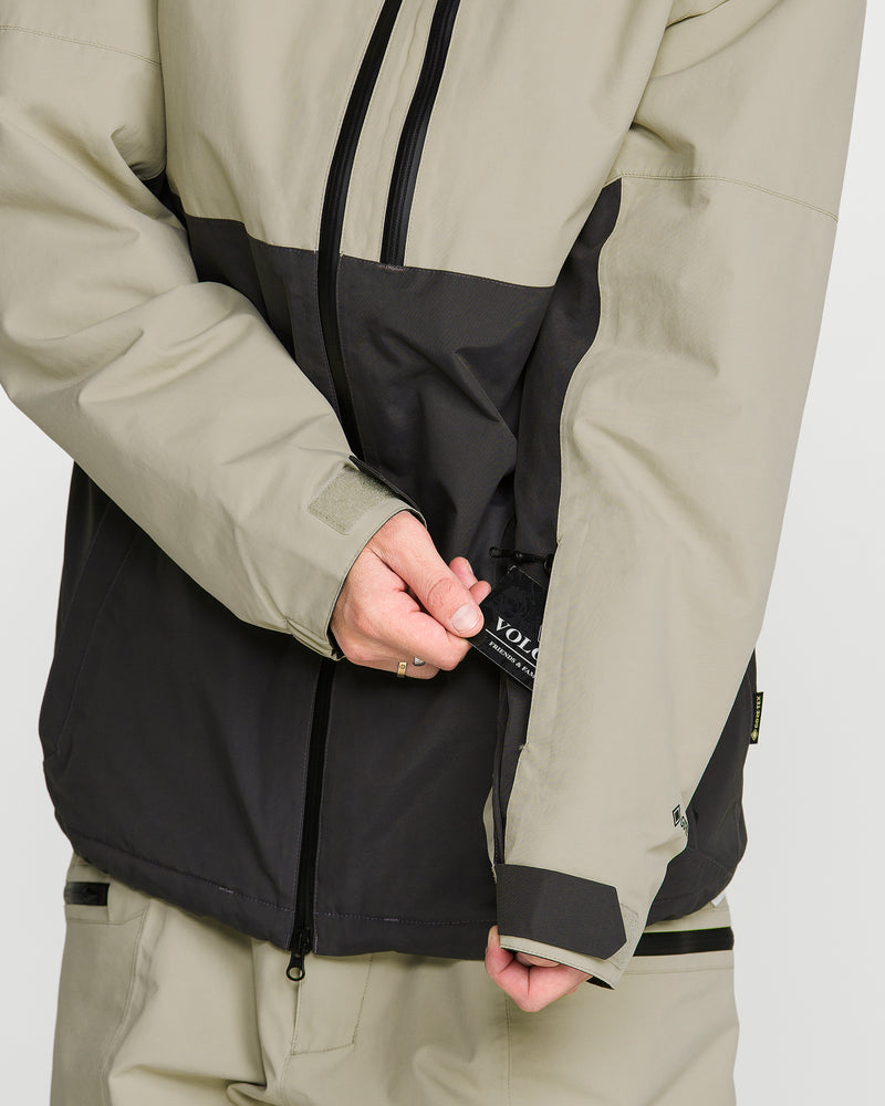 A person wearing the Volcom Men’s L Gore-Tex Jacket in beige and black reaches into a zippered sleeve pocket, revealing a small black item labeled VOL. Only the torso and arms are visible, highlighting the sleek design of this men’s waterproof jacket made from recycled nylon.