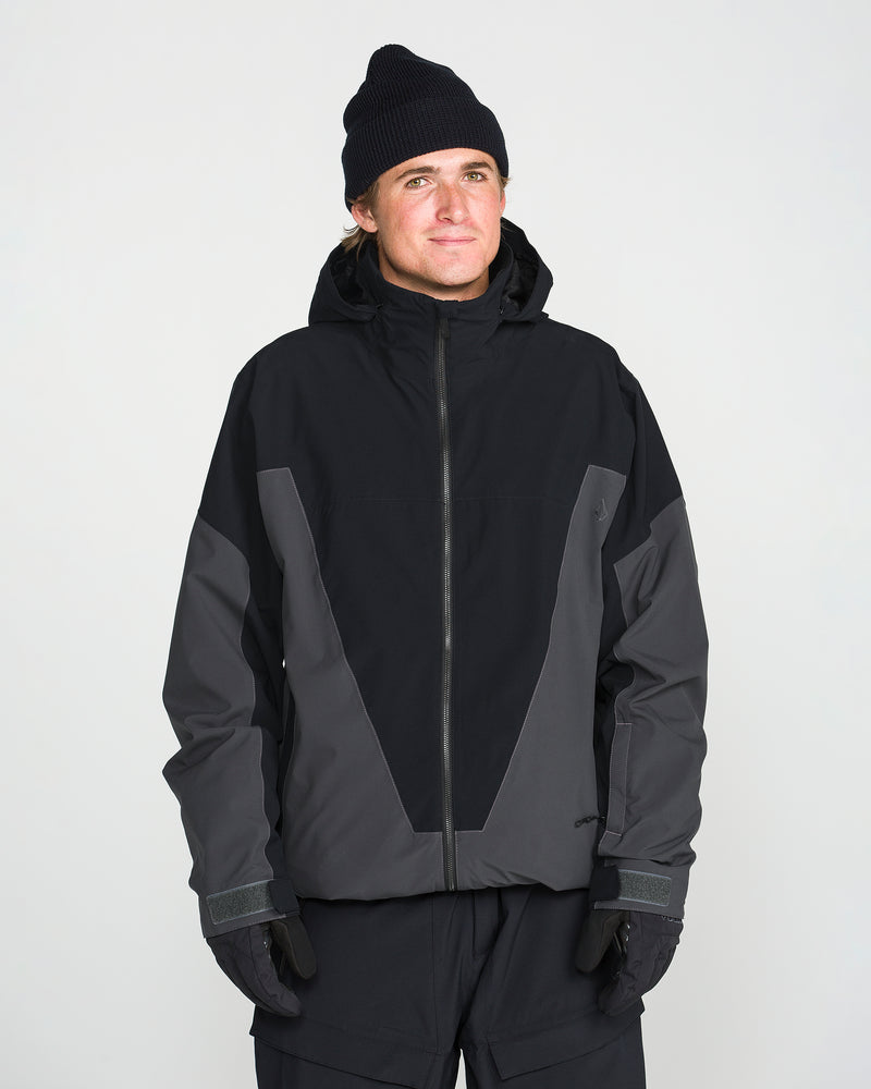 A person wearing a black beanie, black gloves, and dark pants stands against a plain white background, looking slightly upward and smiling softly while showcasing the Volcom Men’s Rnge Stretch 30K Jacket—an outerwear piece crafted from recycled polyester with 4-way stretch in black and gray.