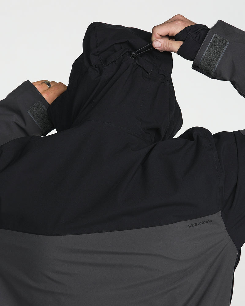 Man adjusts the hood drawstring on a Volcom Men’s Rnge Stretch 30K Jacket, featuring a two-tone design in black and dark gray, crafted from recycled polyester, shown from the back.