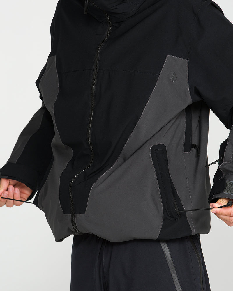 A person adjusts the side drawstrings on a Volcom Men’s Rnge Stretch 30K Jacket in black and gray, with only their torso and hands visible against a plain white background.