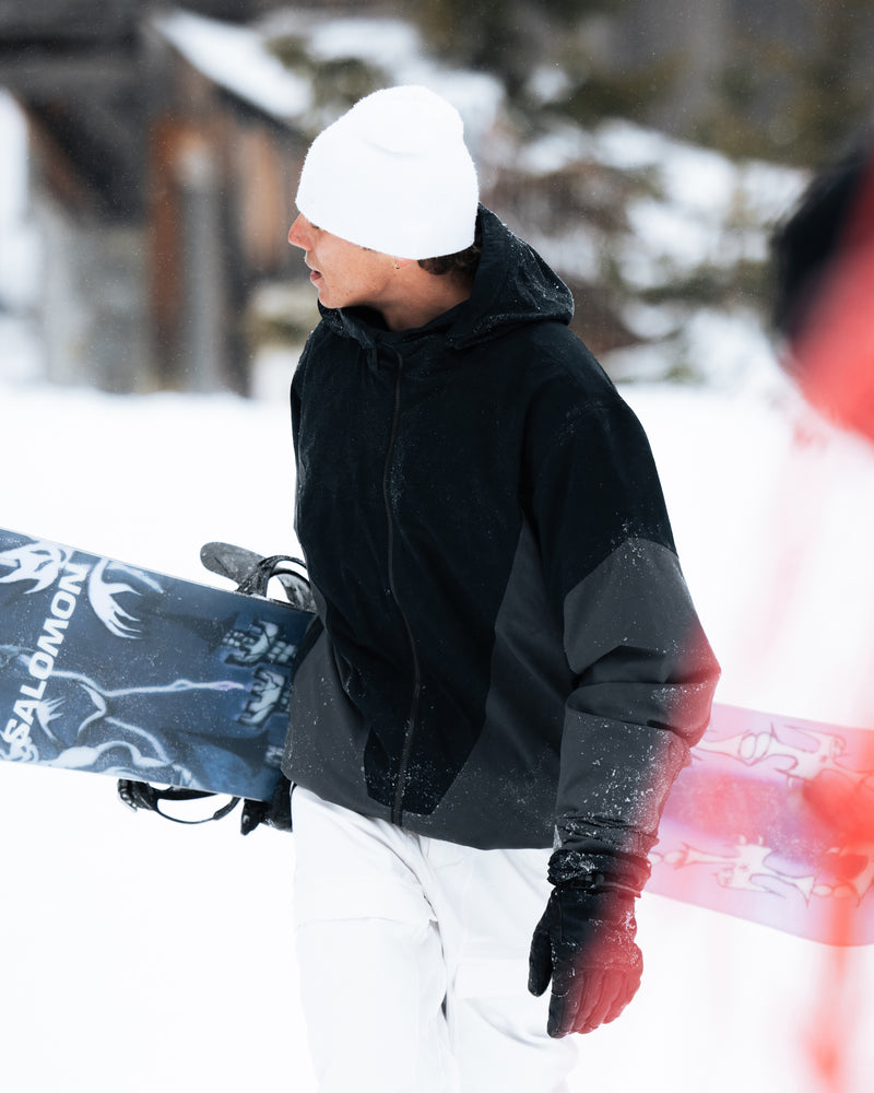 Wearing the Volcom Men’s Rnge Stretch 30K Jacket in black, paired with white pants and a white beanie, a person carries a snowboard through the snow. Trees and a wooden building appear blurred in the background.
