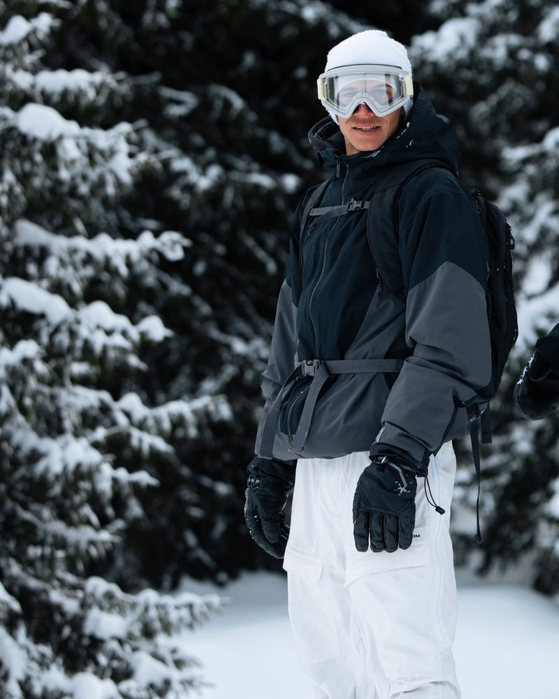Clad in white snow pants, a Volcom Men’s Rnge Stretch 30K Jacket made from recycled polyester, gloves, and ski goggles, a person stands outdoors in a snowy forest, smiling at the camera.