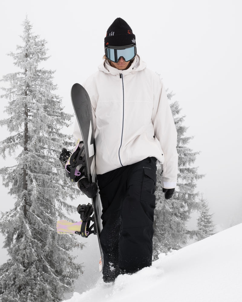 Wearing the Volcom Men’s Rnge Stretch 30K Jacket, black pants, and goggles, a person carries a snowboard while walking up a snowy hill with snow-covered trees in the background.