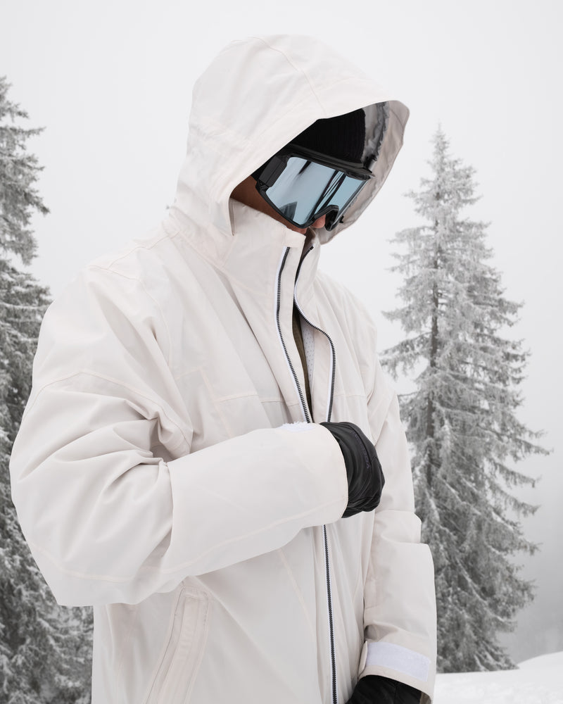Wearing the Volcom Men’s Rnge Stretch 30K Jacket in white, along with black gloves and reflective ski goggles, a person stands outdoors in snowy weather with frosted trees in the background.