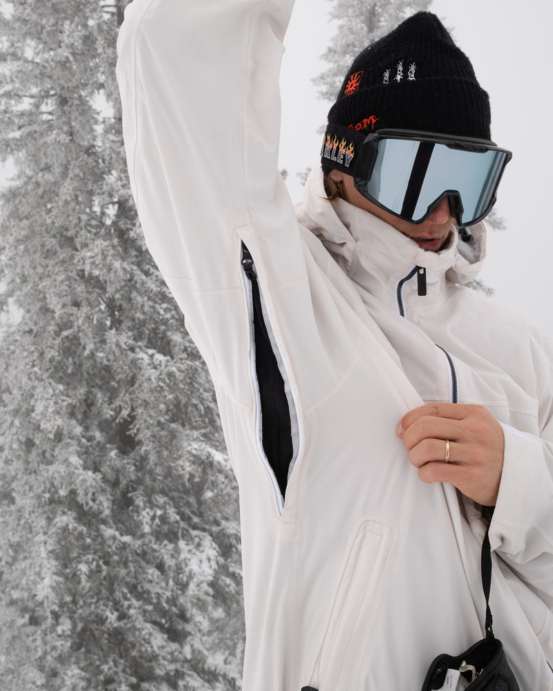 A person wearing the Volcom Men’s Rnge Stretch 30K Jacket in white opens an armpit zipper while outdoors in a snowy, frosty forest. Their face is partially obscured by a beanie and reflective goggles.