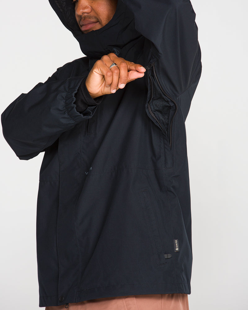 Wearing the Volcom Men’s Dua GORE-TEX Jacket in black, a person unzips the underarm vent to reveal its advanced ventilation system. Their face is partially out of frame, and a ring is visible on their hand.