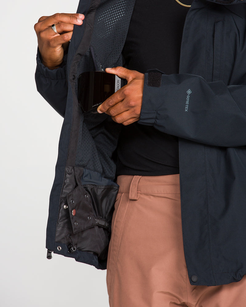 A person places a smartphone into the inner chest pocket of a black Volcom Men’s Dua GORE-TEX Jacket, revealing its mesh lining while wearing tan pants. The innovative Zip Tech feature of this waterproof snow jacket ensures warmth and protection in any weather.